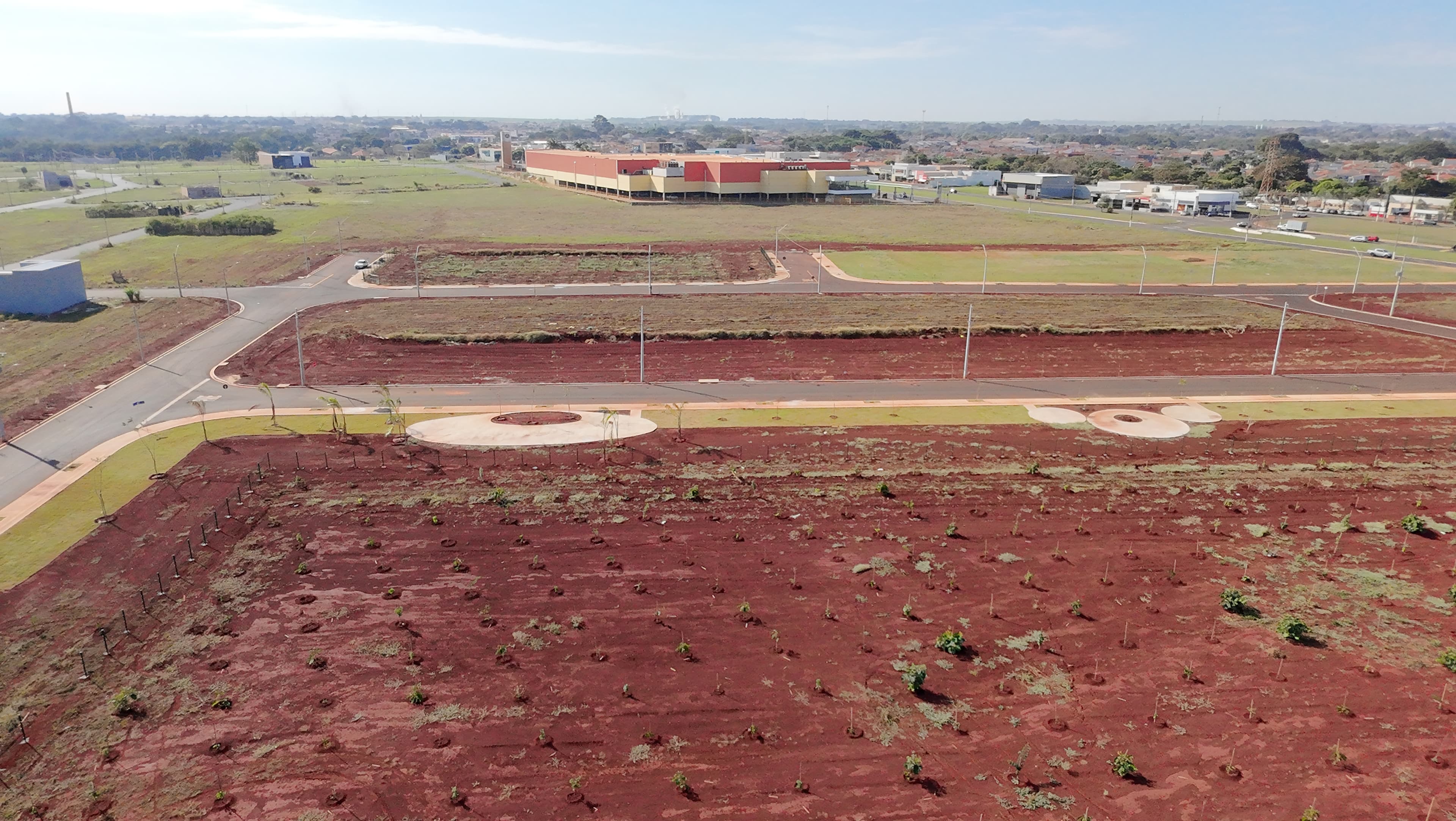 Vista aérea do Bella Verona Boulevard em Maio de 2026, com ruas pavimentadas, mudas plantadas em solo avermelhado e edifício comercial ao fundo